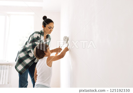 Young woman with kid is painting wall. Charming girl with small child is making repairs in apartment. Mom and toddler apply paint to wall of house with brush. Young woman with kid is painting wall. Charming girl with small child is making repairs in apartment. Mom and toddler apply paint to wall of house with brush. 125192330