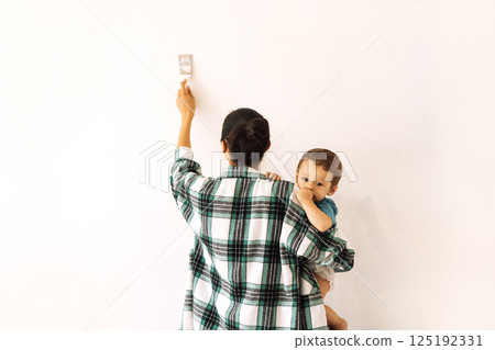 A young woman holds a child and paints a wall. The view from the back. A mother with a small kid is making repairs in an apartment. 125192331