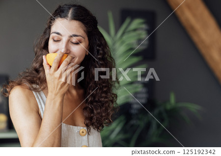 A young attractive woman closes her eyes and sniffs half an orange. A curly-haired smiling girl is eating an orange. A charming brunette enjoys the smell of citrus. 125192342