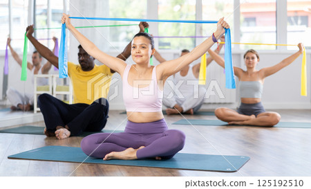 Asian girl conducts Pilates multinational group sitting, elastic band in hands above head body bends 125192510