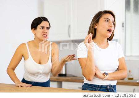 Young Latina dismissively listening to disgruntled girlfriend in home kitchen Young Latina dismissively listening to disgruntled girlfriend in home kitchen 125192588