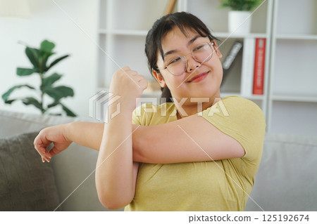 A plump Asian woman is stretching her muscles while sitting on a sofa in her living room at home. 125192674