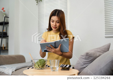Healthy Living and Mindful Eating. A woman takes notes while enjoying a healthy salad at home. Healthy Living and Mindful Eating. A woman takes notes while enjoying a healthy salad at home. 125194268