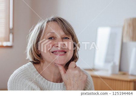 Close-up portrait of an elderly woman in a sweater. A beautiful middle-aged lady in casual clothes is smiling in the living room. A pensioner poses at home. Close-up portrait of an elderly woman in a sweater. A beautiful middle-aged lady in casual clothes is smiling in the living room. A pensioner poses at home. 125194464