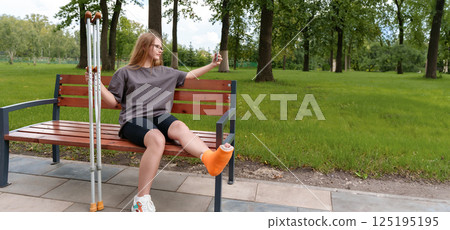 Teenage girl with a cast on her leg seated on a park bench, taking a thoughtful selfie to document the difficulties of her injury and the time she's had to spend in recovery during the summer 125195195