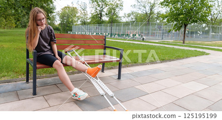 Melancholy teenage girl with a broken leg sits on a park bench, her crutches resting beside her, during a peaceful summer day Melancholy teenage girl with a broken leg sits on a park bench, her crutches resting beside her, during a peaceful summer day 125195199