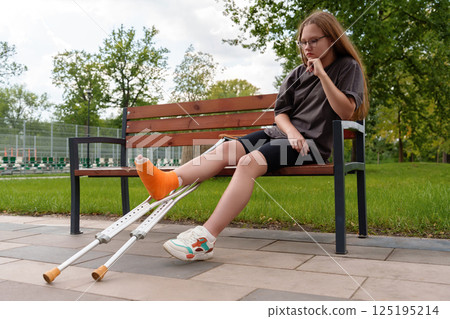 Dejected teen girl with cast on leg sits on park bench, conveying injury frustration and limited mobility, healthcare, insurance, youth rehab 125195214