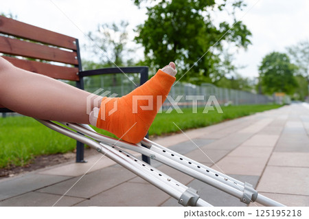 Close-up of cast-encased leg in orange bandage resting on metal crutch, conveying mobility challenges in summer park setting, for healthcare, medical equipment, disability Close-up of cast-encased leg in orange bandage resting on metal crutch, conveying mobility challenges in summer park setting, for healthcare, medical equipment, disability 125195218