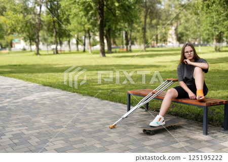 Disheartened girl resting on a park bench with crutches and skateboard nearby 125195222