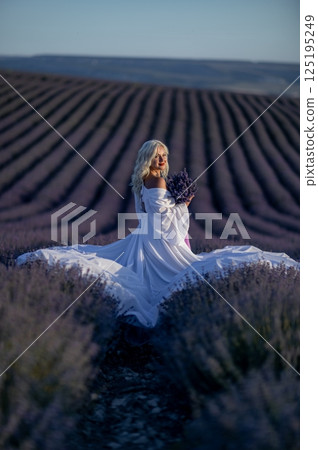 Blonde woman poses in lavender field at sunset. Happy woman in white dress holds lavender bouquet. Aromatherapy concept, lavender oil, photo session in lavender Blonde woman poses in lavender field at sunset. Happy woman in white dress holds lavender bouquet. Aromatherapy concept, lavender oil, photo session in lavender 125195249