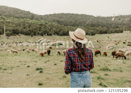 A woman wearing a straw hat stands in a field with a herd of sheep 125195296
