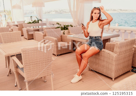 Cafe woman sitting at a table with a cell phone in her hand. She is looking at the screen and she is focused on her phone. The scene takes place in a restaurant or cafe, with multiple chairs. Cafe woman sitting at a table with a cell phone in her hand. She is looking at the screen and she is focused on her phone. The scene takes place in a restaurant or cafe, with multiple chairs. 125195315