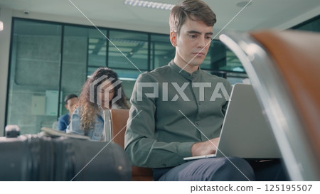 Young handsome man with a suitcase sitting working his laptop at airport terminal hall while waiting landing for her flight, tourist men in international airport, travel trip Young handsome man with a suitcase sitting working his laptop at airport terminal hall while waiting landing for her flight, tourist men in international airport, travel trip 125195507