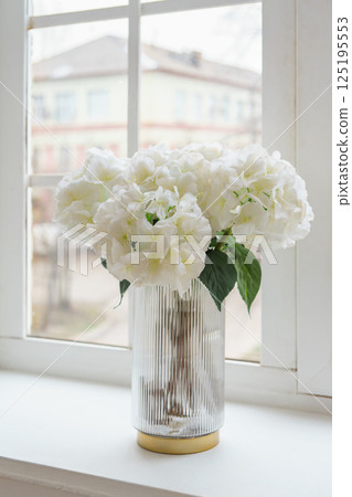 Bouquet of white artificial decorative hydrangea, hortensia in vase on windowsill in living room 125195553
