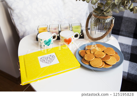 Cozy breakfast scene with mini pancakes, drinks, and a yellow album on a white table 125195828