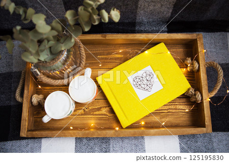 Cozy autumn scene with yellow book, mugs, and eucalyptus in a rustic wooden tray, decorated with fairy lights 125195830