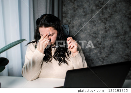 Tired woman working at home with laptop holding eyeglasses rubbing eyes and feeling fatigue. Stress and frustration concept. Tired eyes Tired woman working at home with laptop holding eyeglasses rubbing eyes and feeling fatigue. Stress and frustration concept. Tired eyes 125195835