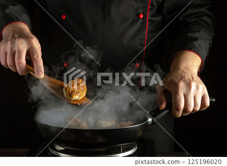 A skilled chef prepares meatballs in a frying pan, surrounded by steam and smoke. The warm lighting enhances the ambiance as the chef focuses on the cooking process 125196020