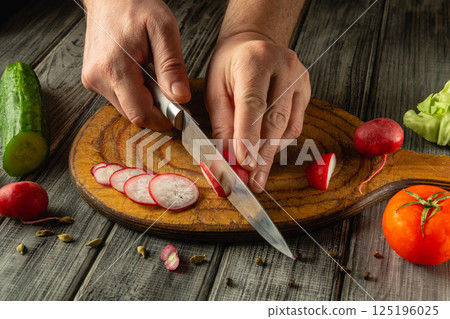 Hands skillfully slice radishes into thin rounds on a wooden cutting board. Nearby, fresh cucumbers, tomatoes, and lettuce add vibrant colors to this culinary preparation in a cozy kitchen Hands skillfully slice radishes into thin rounds on a wooden cutting board. Nearby, fresh cucumbers, tomatoes, and lettuce add vibrant colors to this culinary preparation in a cozy kitchen 125196025