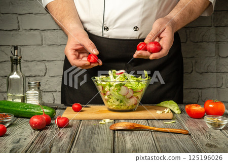 A chef in a black apron prepares a fresh salad by adding radishes to a bowl of mixed greens on a rustic wooden table in a well-lit kitchen A chef in a black apron prepares a fresh salad by adding radishes to a bowl of mixed greens on a rustic wooden table in a well-lit kitchen 125196026