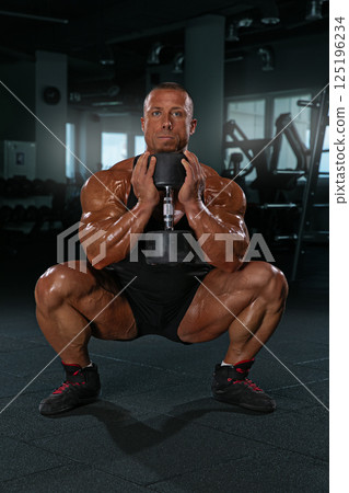 high level athlete focuses on strength training by squatting with a dumbbell in an empty gym. The strong physique and dedication highlight an elite fitness routine in the evening. 125196234