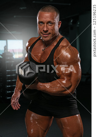 high level athlete poses with a dumbbell in a well equipped gym. The individual exhibits impressive muscle definition and a focused expression. high level athlete poses with a dumbbell in a well equipped gym. The individual exhibits impressive muscle definition and a focused expression. 125196235