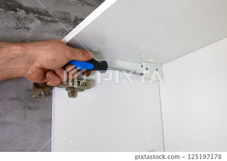 A man uses a screwdriver to fix a bracket mounted on a white rack, a cabinet mounted on the wall.  125197178