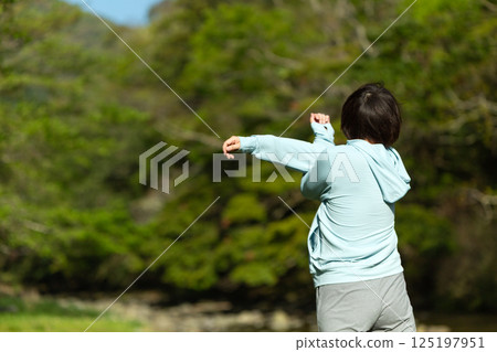 A middle-aged woman doing exercises by the riverside in the fresh greenery A middle-aged woman doing exercises by the riverside in the fresh greenery 125197951