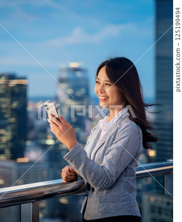 Smiling young businesswoman using her smartphone on a rooftop with a view of the city's skyline 125199494