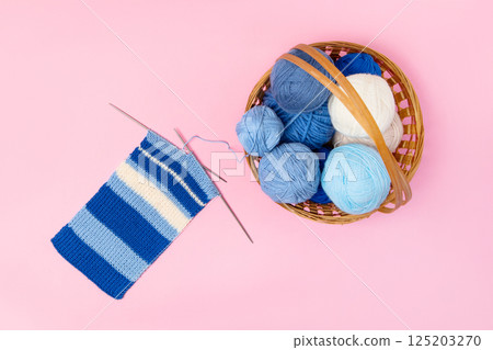 Blue and beige knitting balls in basket with knitting needles and knitted scarf on pink background. Set for the hobby of knitting warm clothes or toys. Concept needlework 125203270