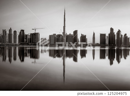 Monochrome city skyline with tall skyscrapers reflecting on calm water under a cloudy sky, Dubai Marina bay UAE 125204975
