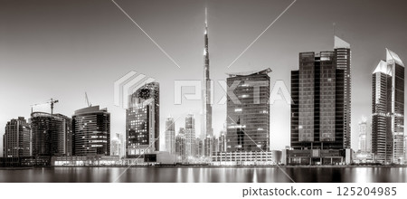 Monochrome city skyline with tall skyscrapers reflecting on calm water under a cloudy sky, Dubai Marina bay UAE 125204985