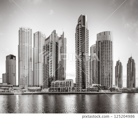 Monochrome city skyline with tall skyscrapers reflecting on calm water under a cloudy sky, Dubai Marina bay UAE 125204986