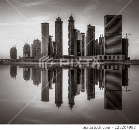 Monochrome city skyline with tall skyscrapers reflecting on calm water under a cloudy sky, Dubai Marina bay UAE 125204998