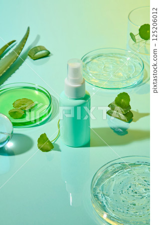 Vertical photo of a plastic cosmetic spray bottle with a turquoise packaging. Centella leaves placed in a petri dish containing green liquid. Other petri dishes contain gel extracted from aloe vera. Vertical photo of a plastic cosmetic spray bottle with a turquoise packaging. Centella leaves placed in a petri dish containing green liquid. Other petri dishes contain gel extracted from aloe vera. 125206012