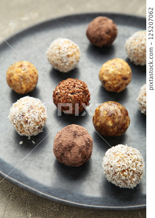 Healthy raw energy balls. Candy vegan balls of dates, nuts and coconut on plate on grey background. Top view, concept of useful home-made candies without sugar. Vertical photo Healthy raw energy balls. Candy vegan balls of dates, nuts and coconut on plate on grey background. Top view, concept of useful home-made candies without sugar. Vertical photo 125207062