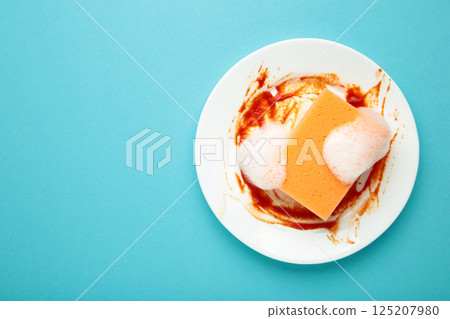 White round dirty plate with red sauce, orange foam sponge cleaning dish on blue background. 125207980