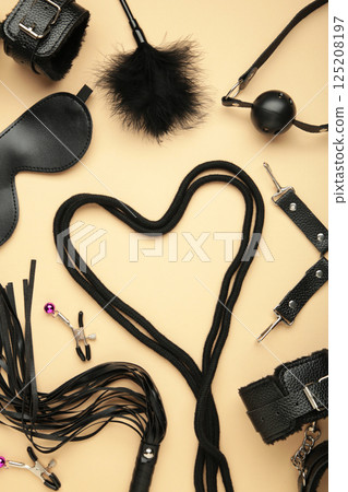 BDSM set on beige background. BDSM accessories and rope in the shape of a heart. Mask, handcuffs, ball gag and a whip. Vertical photo 125208197