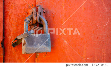 Rusty padlock on bright orange metal door with weathered surface texture 125210389