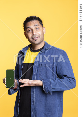 Indian content creator presenting green screen on mobile device, standing against orange background in the studio. Young male model wearing casual clothes and a man bun, internet channel. 125211564