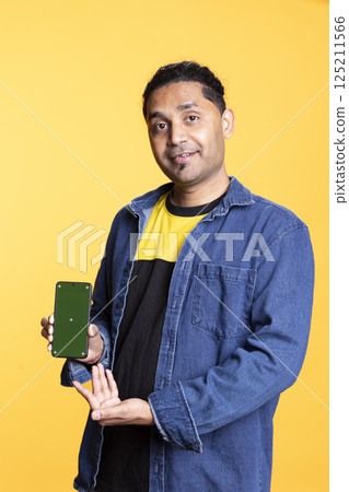 Smiling indian adult presenting mobile phone with isolated mockup screen, staying against studio background. Young casual guy creating a promotion with blank display on gadget. 125211566