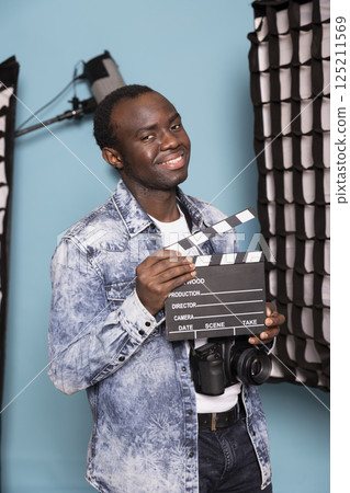 Portrait of smiling african american man standing in studio, holding clapperboard, ready for a shoot. Black male photographer in denim jacket grasping a film slate, standing among lighting equipment. 125211569