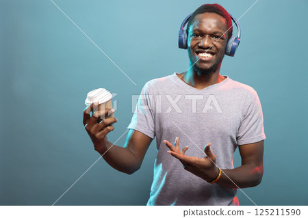 Cheerful young guy gesturing to his coffee cup and looking at camera. Portrait of black male person standing with a hot drink and enjoying music through his wireless headphones. Cheerful young guy gesturing to his coffee cup and looking at camera. Portrait of black male person standing with a hot drink and enjoying music through his wireless headphones. 125211590