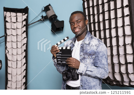 African american filmmaker standing in studio surrounded by lighting equipment, holding a clapperboard. Portrait of black male photographer with camera hanging over his neck and film slate. African american filmmaker standing in studio surrounded by lighting equipment, holding a clapperboard. Portrait of black male photographer with camera hanging over his neck and film slate. 125211591