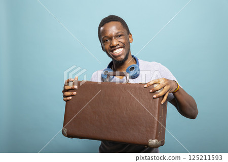 Cheerful african american man with a suitcase and headphones, excited for his journey. Black male tourist looking at the camera and posing with his luggage against an isolated background. 125211593