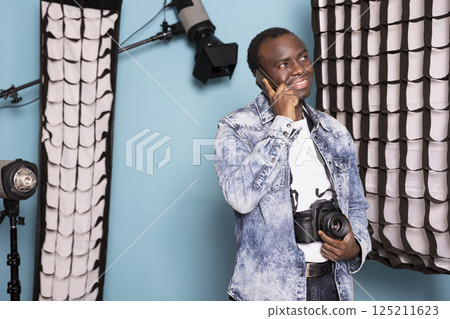 African american man grins widely while speaking on his mobile, holding his dslr device with one hand. Joyful male photographer with a camera around his neck stands, having a phone call in studio. African american man grins widely while speaking on his mobile, holding his dslr device with one hand. Joyful male photographer with a camera around his neck stands, having a phone call in studio. 125211623