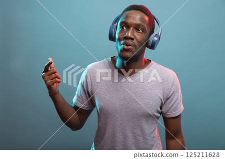 Black man wearing wireless headset, moving to the music with excitement, set against an isolated backdrop. Happy male model dancing with bluetooth headphones, enjoying upbeat tunes. Black man wearing wireless headset, moving to the music with excitement, set against an isolated backdrop. Happy male model dancing with bluetooth headphones, enjoying upbeat tunes. 125211628