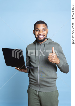 African american young guy typing on his laptop against blue background, composing emails and working on paperwork. Male overjoyed person multitasking in studio, cheerful adult. African american young guy typing on his laptop against blue background, composing emails and working on paperwork. Male overjoyed person multitasking in studio, cheerful adult. 125211633