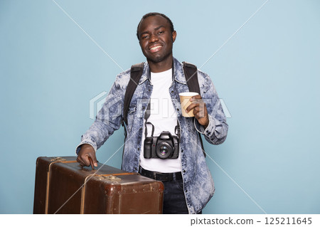 African american photographer dressed in a denim jacket carrying his luggage and backpack preparing for a journey. He holds a camera for capturing moments and a takeaway coffee for the road. 125211645