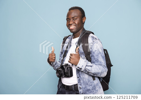 Cheerful african american photographer making an enthusiastic thumbs up gesture, ready to embark on his next creative journey. He smiles brightly while carrying his backpack and DSLR camera. 125211709